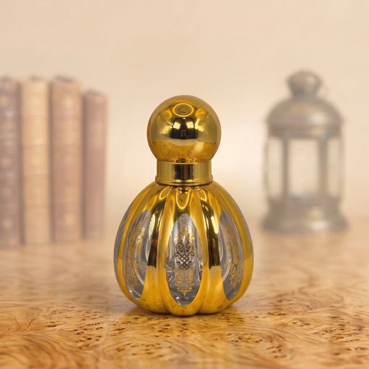 A small ornate gold perfume bottle with a spherical cap and ribbed body, standing on a wooden surface with a softly blurred background featuring books and a lantern.