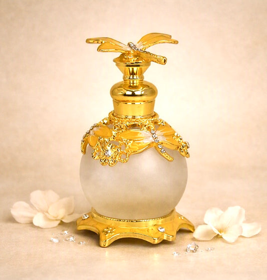An ornate perfume bottle with a round milky white body, decorated with shiny gold metal detailing, a gold floral cap, and a gold pedestal base, styled with white flower petals on a beige background.