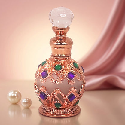 An ornate decorative perfume bottle with a rose gold metallic body, a faceted clear glass stopper, and purple and green jewel accents, set against a soft pink satin drape background with two pearl-like beads on the left.