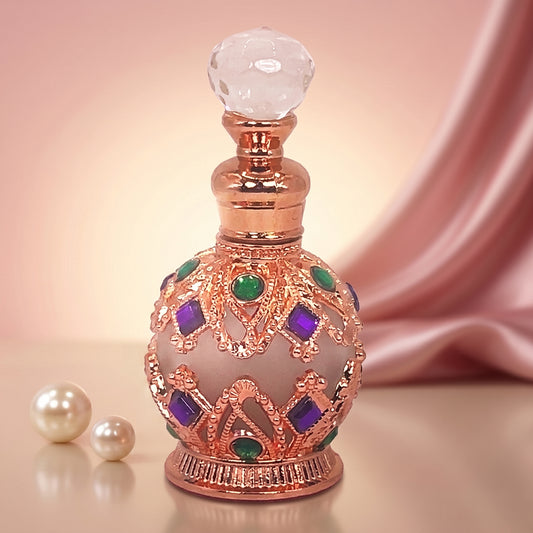 An ornate decorative perfume bottle with a rose gold metallic body, a faceted clear glass stopper, and purple and green jewel accents, set against a soft pink satin drape background with two pearl-like beads on the left.